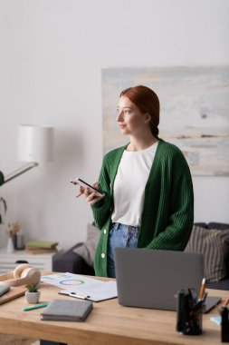 Canlı kızıl saçlı genç bir kadın modern dairesinde telefonuyla uğraşırken rahatlıyor..