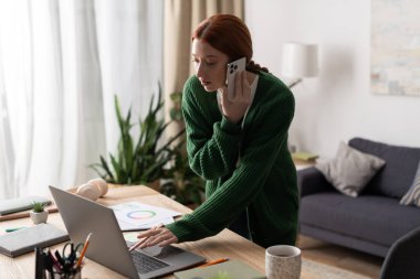 Kızıl saçlı genç bir kadın evde telefonla konuşurken dizüstü bilgisayarına odaklanıyor..