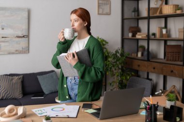 Kızıl saçlı genç bir kadın modern dairesinde, elinde fincanla rahatlıyor ve gününü planlıyor..