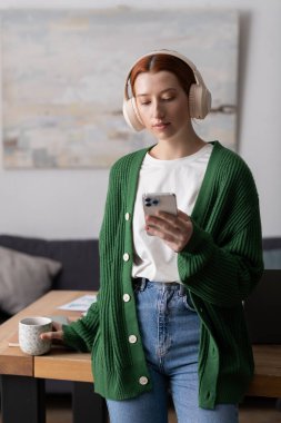 Modern bir dairede, kızıl saçlı genç bir kadın sıcak bir içeceği yudumlarken telefonunu kontrol eder..