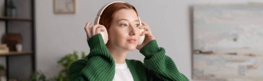 Kızıl saçlı kadın, günlük giysiler içinde evde dinlenirken müzik dinliyor, afiş.