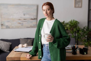 Parlak kızıl saçlı kadın, elinde içki tutarken şık dairesinde rahat bir anın tadını çıkarıyor..