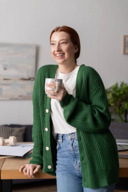 Kırmızı saçlı neşeli genç bir kadın şık dairesinde dinlenirken çayını yudumluyor..