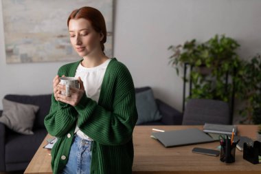 Kızıl saçlı genç bir kadın şık bir dairede sıcak içkisinin tadını çıkarıyor..
