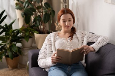 Kızıl saçlı genç bir kadın modern dairesinde rahatça bir kitap okuyor..