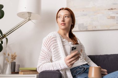 Kızıl saçlı genç bir kadın telefonunu tutarak evde vakit geçirmekten zevk alıyor..