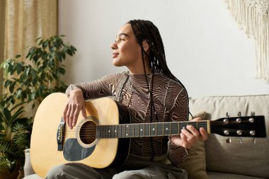 Örgülü genç bir Afrikalı Amerikalı kadın modern, rahat dairesinde gitar çalıyor..