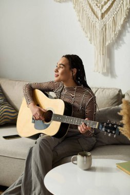 Saçları örgülü genç bir Afrikalı Amerikalı kadın şık, rahat bir dairede gitarını tıngırdatmaktan hoşlanıyor..