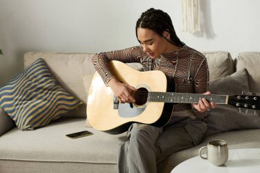 Saçları örgülü genç bir Afrikalı Amerikalı kadın şık dairesinde gitar çalıyor, neşe saçıyor..