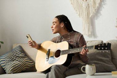 Genç bir Afrikalı Amerikalı kadın şık bir dairede telefonuna bakarken gitar çalıyor..