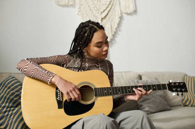 Saçları örgülü genç bir kadın şık dairesinde gitar çalıyor, müziğine dalmış..