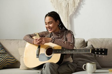 Saçları örgülü genç bir zenci kadın evde gitar çalarken telefonunu kullanarak gülümsüyor..