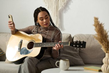 Genç bir Afrikalı Amerikalı kadın rahat bir yerde telefonunu kontrol ederken koltuğunda gitar çalıyor..