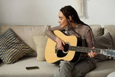 Örgülü genç siyahi bir kadın rahat, modern dairesinde gitar çalıyor, müzikten hoşlanıyor..