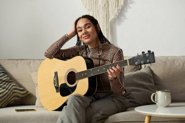 Saçları örgülü genç bir Afrikalı Amerikalı kadın rahat dairesinde gitar çalıyor, neşe saçıyor..