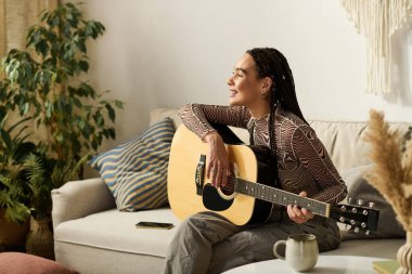 Genç siyahi bir kadın, modern bir dairede gitar çalarak kanepede dinleniyor..