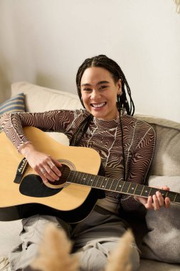 Genç bir Afrikalı Amerikalı kadın modern, rahat dairesinde gitar çalarken gülümsüyor..