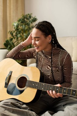 Saçları örgülü genç bir Afrikalı Amerikalı kadın rahat dairesinde otururken gitar çalmaktan hoşlanıyor..