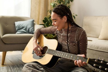 Örgülü genç bir Afrikalı Amerikalı kadın modern dairesinde gülümserken gitar çalıyor..