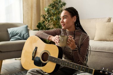 Saçları örgülü genç siyah bir kadın yerde dinleniyor, çay içiyor ve gitar çalıyor..