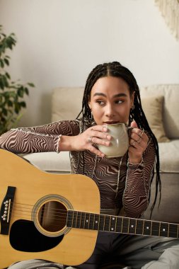 Genç bir Afrikalı Amerikalı kadın, dairesinde gitarını tıngırdatırken kahve yudumluyor..