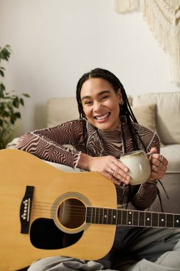 Saçları örgülü, gitarının yanında kupa tutan genç bir zenci kadın..