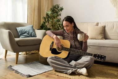 Saçları örgülü, elinde kupa olan ve evde gitar çalan genç bir zenci kadın..