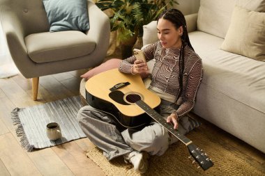 Genç bir kadın elinde gitarıyla telefonuyla yerde rahatça oturuyor..