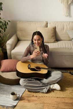 Genç bir Afrikalı Amerikalı kadın şık dairesinde dinlenirken gitar çalıyor ve gülümsüyor..