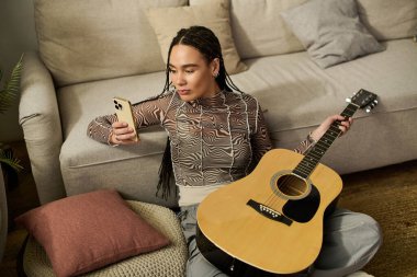Örgülü genç bir kadın, elinde gitar tutarken telefonunu kontrol ederken yerde rahatça oturuyor..