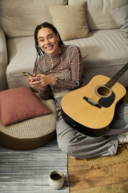 Genç bir Afrikalı Amerikalı kadın yerde oturuyor, elinde gitar ve telefon ile gülümsüyor..