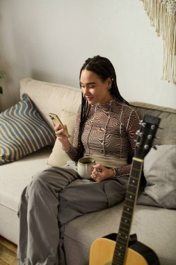 Saçları örgülü genç siyah bir kadın şık kanepesinde dinleniyor, çayını yudumluyor ve telefonunu kullanıyor..