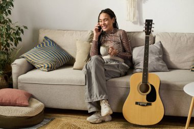 Örgülü genç bir kadın telefonda konuşurken kanepede gülüyor ve çay içiyor..