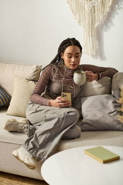 Rahat bir dairede, genç bir Afrikalı Amerikalı kadın telefonunu kullanırken kahveyle rahatlıyor..