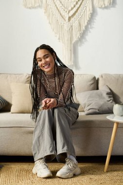 Genç Afrikalı Amerikalı bir kadın şık örgüleriyle modern dairesinde rahatlıyor, sıcak bir şekilde gülümsüyor..