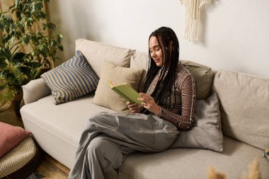 Sıcak bir dairede, genç bir Afrikalı Amerikalı kadın yeşil bir kitap okurken rahatlıyor..