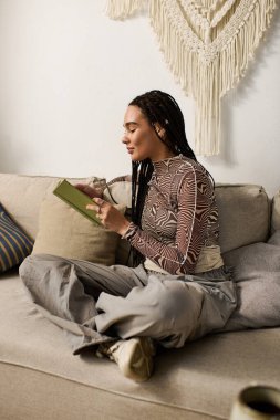 Saçları örgülü genç bir Afrikalı Amerikalı kadın rahat rahat evinde oturup kitap okuyor..