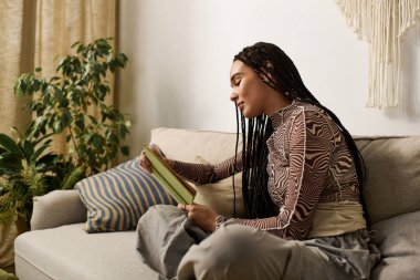 Örgülü genç bir kadın modern dairesinde rahatça oturup kitabına odaklanıyor..