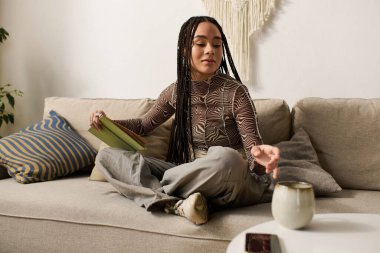 Saçları örgülü genç siyahi bir kadın kitap okuyarak rahat bir şekilde dairesinde oturuyor.