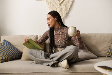 Genç bir Afrikalı Amerikalı kadın koltuğunda oturup elinde sıcak bir içecekle kitap okuyor..