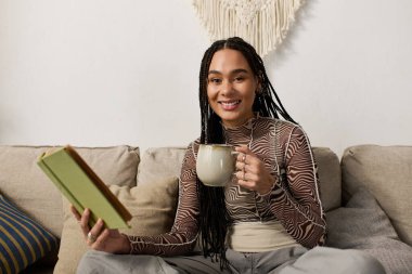 Genç bir Afrikalı Amerikalı kadın şık dairesinde rahatça oturur, çay eşliğinde kitabın tadını çıkarır..