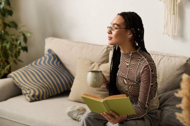 Genç bir Afrikalı Amerikalı kadın şık dairesinde kitap ve çayla dinleniyor..