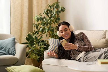 Örgülü genç bir kadın kanepede dinleniyor, telefonunda müzik dinlerken gülümsüyor..