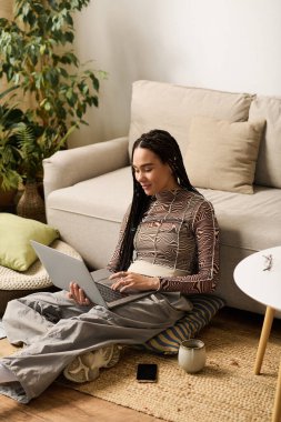 Genç bir kadın şık dairesinde yerde oturuyor, rahat bir şekilde dizüstü bilgisayarıyla çalışıyor..