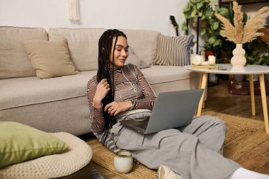 Genç Afrikalı Amerikalı bir kadın lüks dairesinde rahat bir şekilde oturur ve dizüstü bilgisayarına odaklanır..