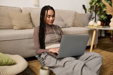 Saçları örgülü genç siyah bir kadın, şık bir dairede dizüstü bilgisayarına odaklanmış bir şekilde yerde oturuyor..