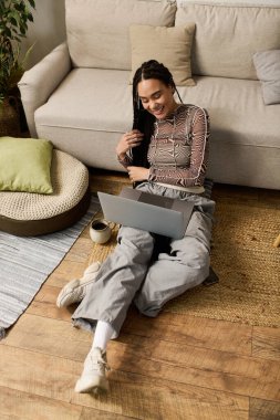 Parlak ve şık genç bir kadın, rahat bir dairede dizüstü bilgisayarıyla yerde oturuyor..