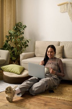 Genç siyahi bir kadın dizüstü bilgisayarı ve sıcak içkisiyle evinde dinleniyor..