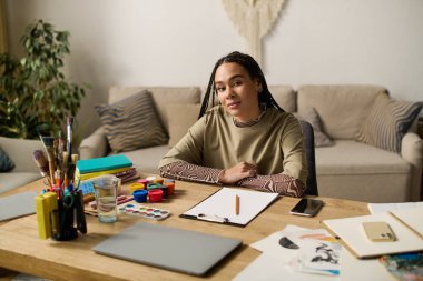Genç bir Afrikalı Amerikalı kadın, sanat malzemeleriyle çevrili modern dairesinde güven içinde oturuyor..