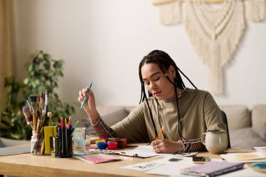 Örgülü genç bir kadın renkli malzemelerle çevrili modern dairesinde sanat yaratıyor..